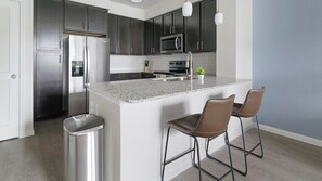 A modern kitchen with dark cabinets, stainless steel appliances, a granite countertop, and two bar stools. - Landing | Beautiful 1BD, Gym (Conroe)
