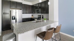 A modern kitchen with dark cabinets, stainless appliances, a granite countertop, and bar stools. - Landing | Stylish 1BD, Gym (Conroe)