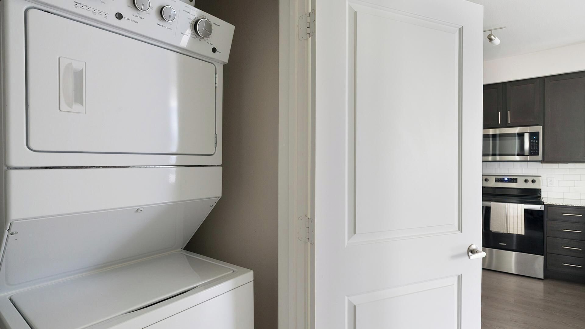 A laundry area with stacked washers and dryers, a white door, and a kitchen with dark cabinets and stainless steel appliances.