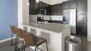 A modern kitchen with dark cabinets, stainless steel appliances, a granite countertop, and three bar stools. - Landing | Chic 1BD, Gym (Conroe)