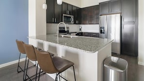 A modern kitchen with dark cabinets, stainless steel appliances, a granite countertop, and three bar stools. - Landing | Modern 1BD, Gym (Conroe)