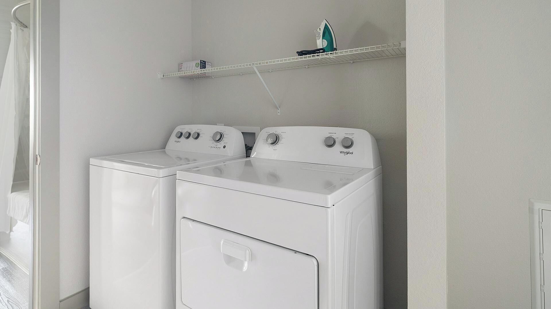 Un lavadero con dos lavadoras y secadoras Whirlpool blancas, un estante metálico arriba y una puerta blanca a la izquierda.