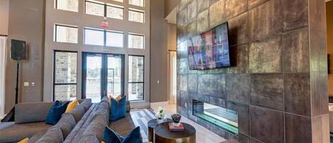 A spacious lobby with a large seating area, a flat-screen TV mounted on a textured wall, and large windows allowing natural light.