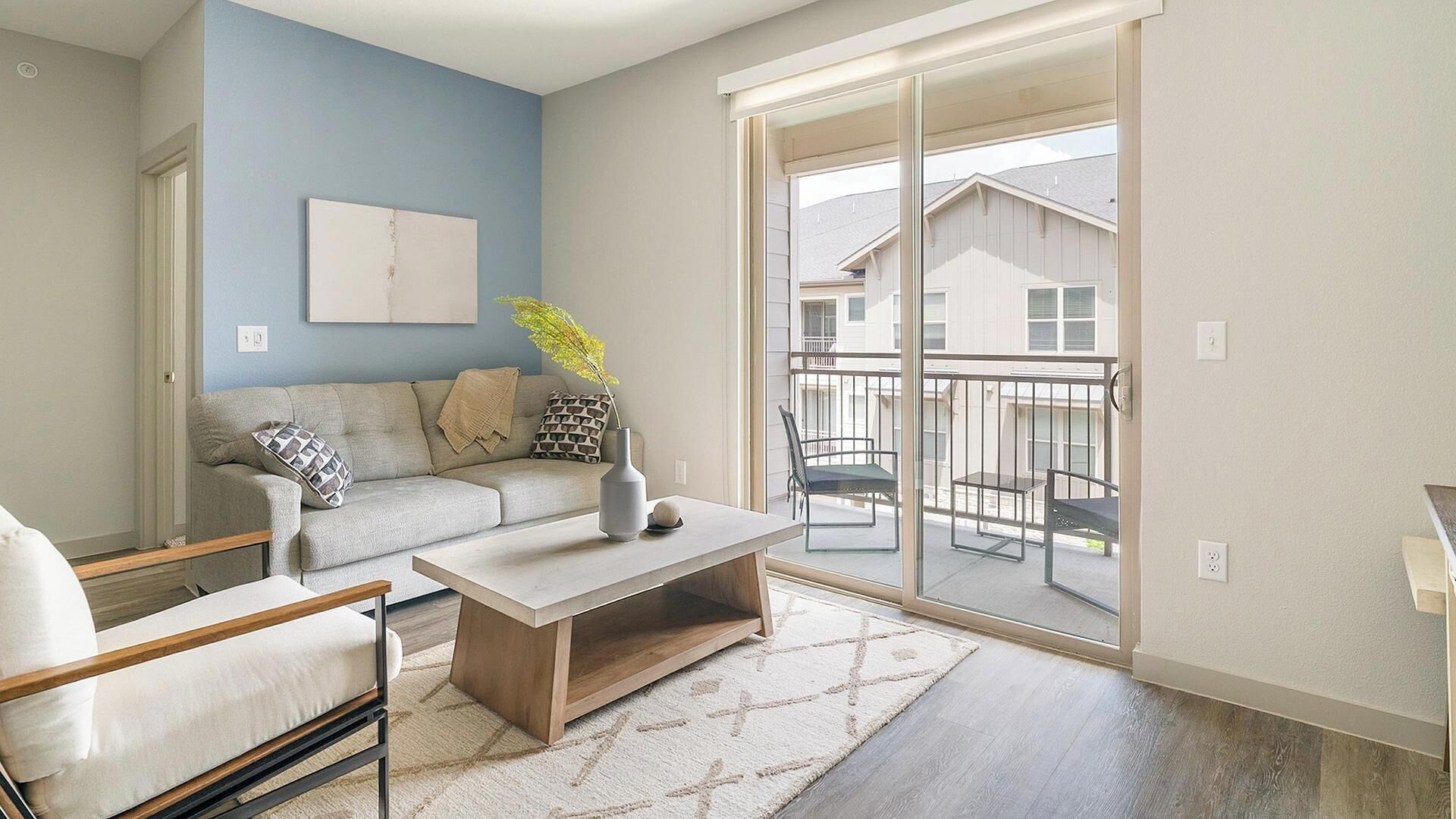 A modern apartment with a sofa, coffee table, and chairs, a balcony with a view of houses, and a blue accent wall.