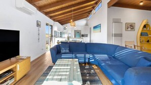 Ein modernes Wohnzimmer mit einem großen, blauen Ecksofa, einem Flachbildfernseher, Holzböden und einer Küche im Hintergrund.