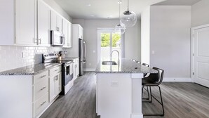 A modern kitchen with white cabinets, a granite countertop, stainless steel appliances, and a black chair. - Landing | Beautiful 2BD, Gym (Longs)