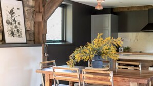 Un comedor con sillas de madera y una mesa, un florero con flores amarillas, una ilustración botánica enmarcada y una ventana con vistas.