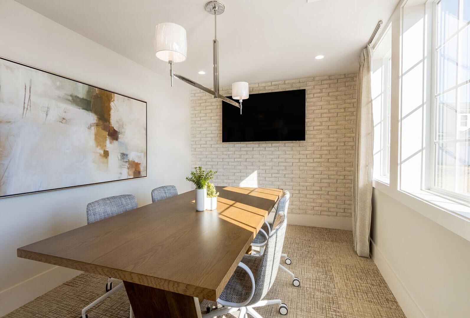 A conference room with a long table, chairs, a potted plant, a mounted TV, and a large abstract painting on the wall.
