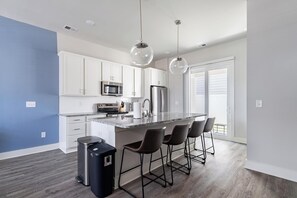 A modern kitchen with white cabinets, a central island with bar stools, stainless steel appliances, and pendant lighting. - Landing | Stunning 2BD, Gym (Longs)