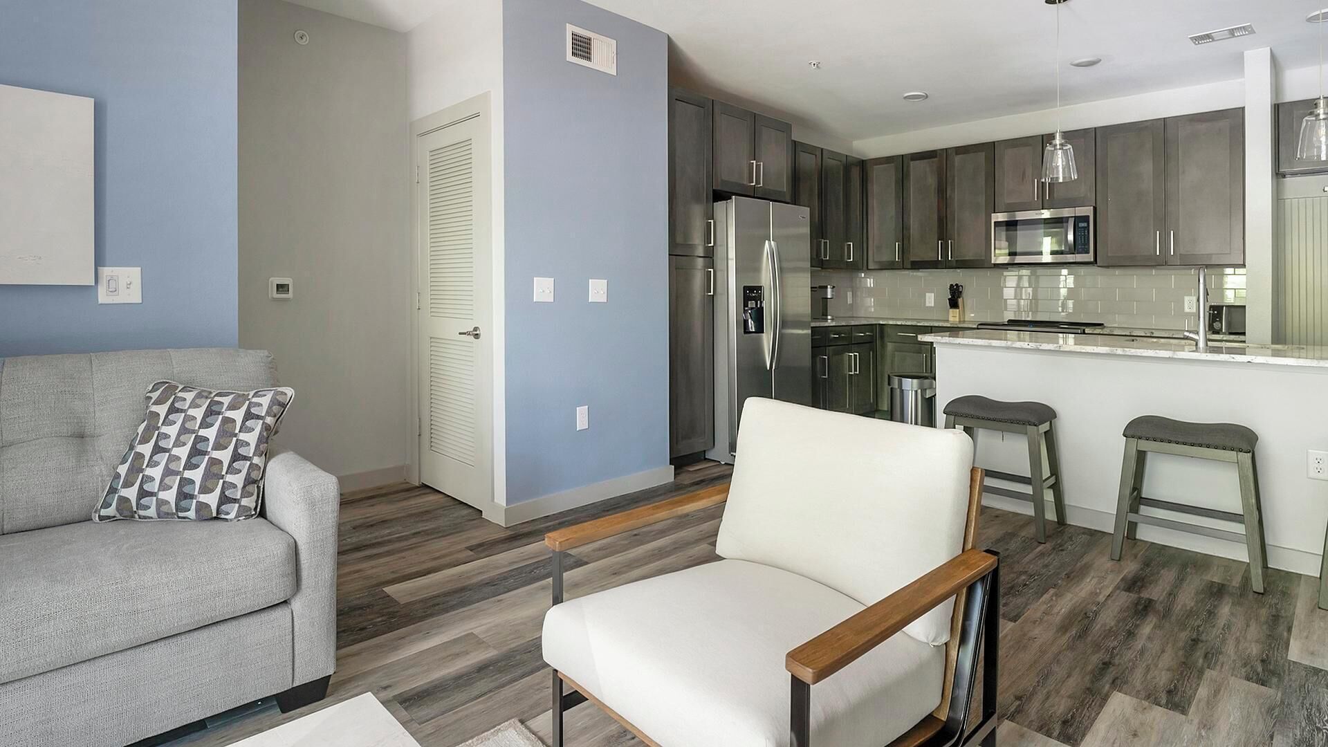 A modern living room with a grey sofa, a white armchair, and a patterned throw pillow. A wooden floor and a kitchen with grey cabinets and stainless steel appliances are visible.
