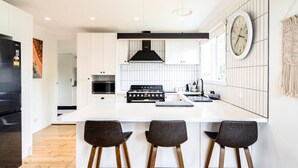 Une cuisine moderne dotée d’un îlot central, d’une cuisinière, d’un four et d’un four à micro-ondes. On y trouve trois tabourets de bar et une horloge au mur.