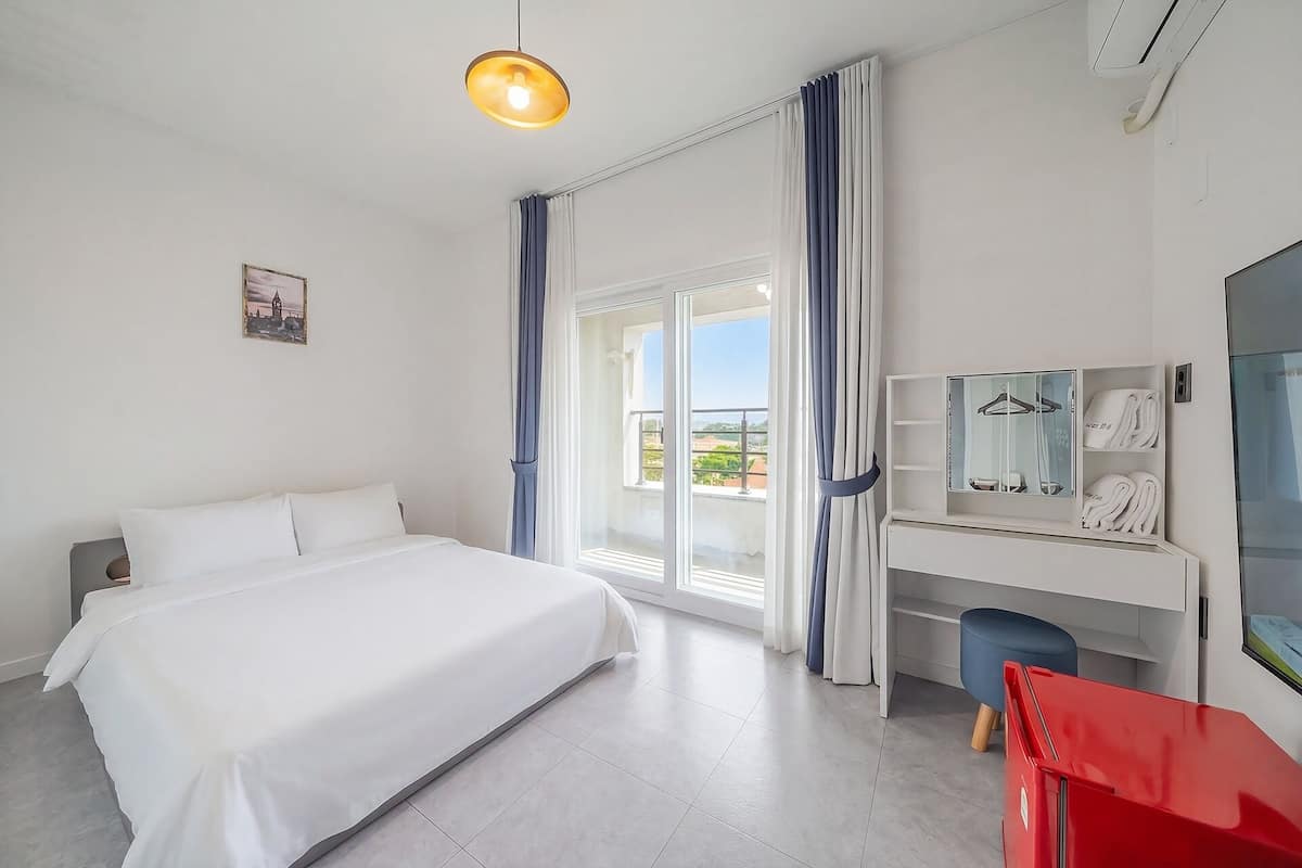 Habitación de hotel con una cama, un escritorio con espejo, una silla roja y un ventanal con cortinas.