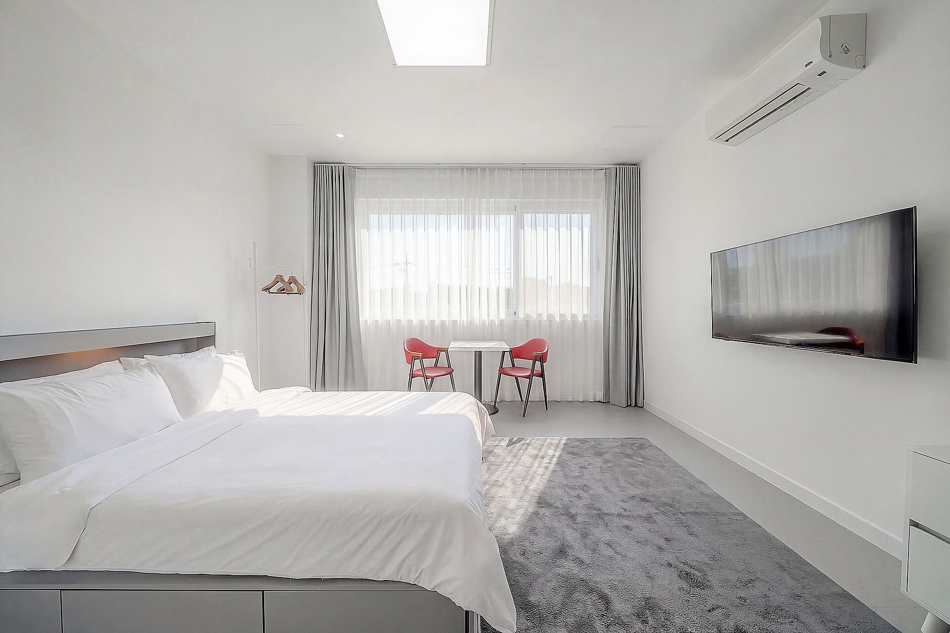 A hotel room with a bed, a TV mounted on the wall, a small table with two chairs, and a window with curtains.