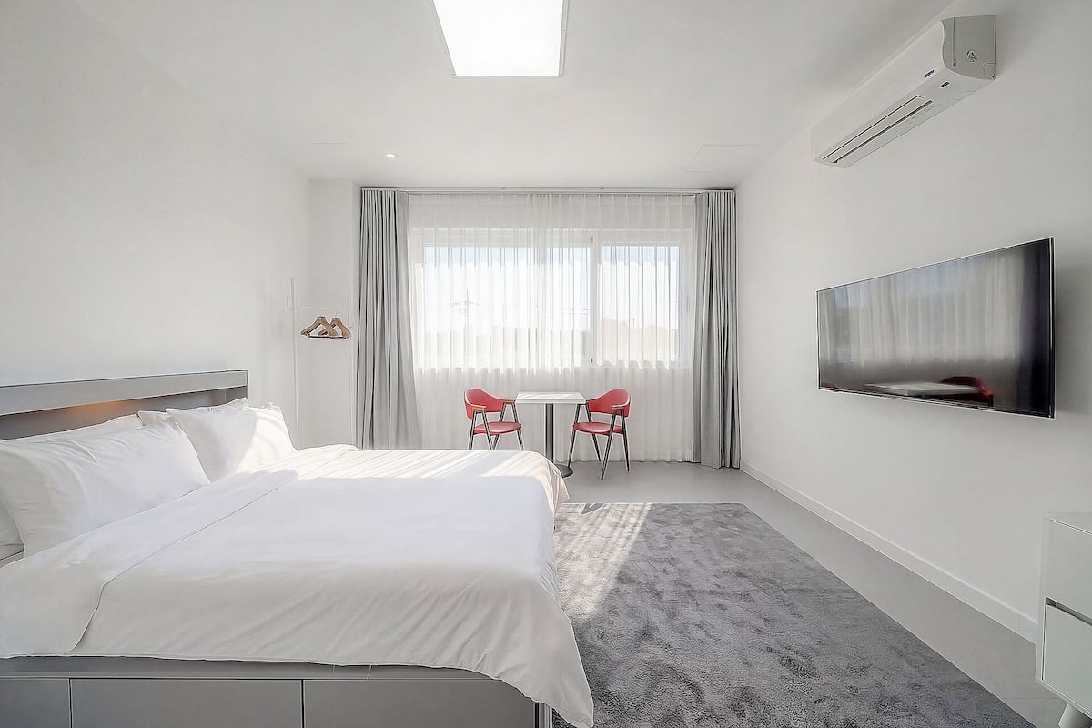 Habitación de hotel con cama, televisión montada en la pared, una mesita con dos sillas y una ventana con cortinas.