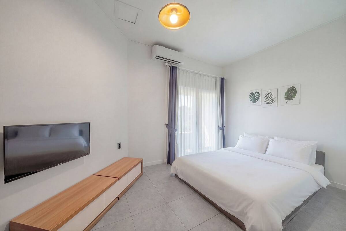 Un dormitorio con cama, televisión, banca y lámpara de techo.