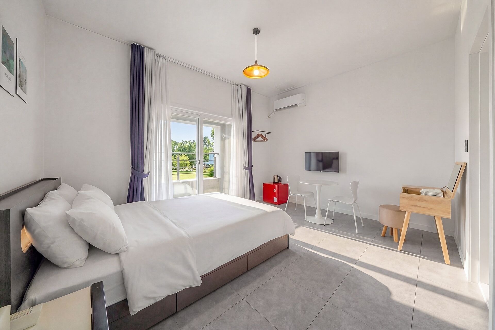 A modern bedroom with a bed, a TV, a desk, and a view of greenery outside.