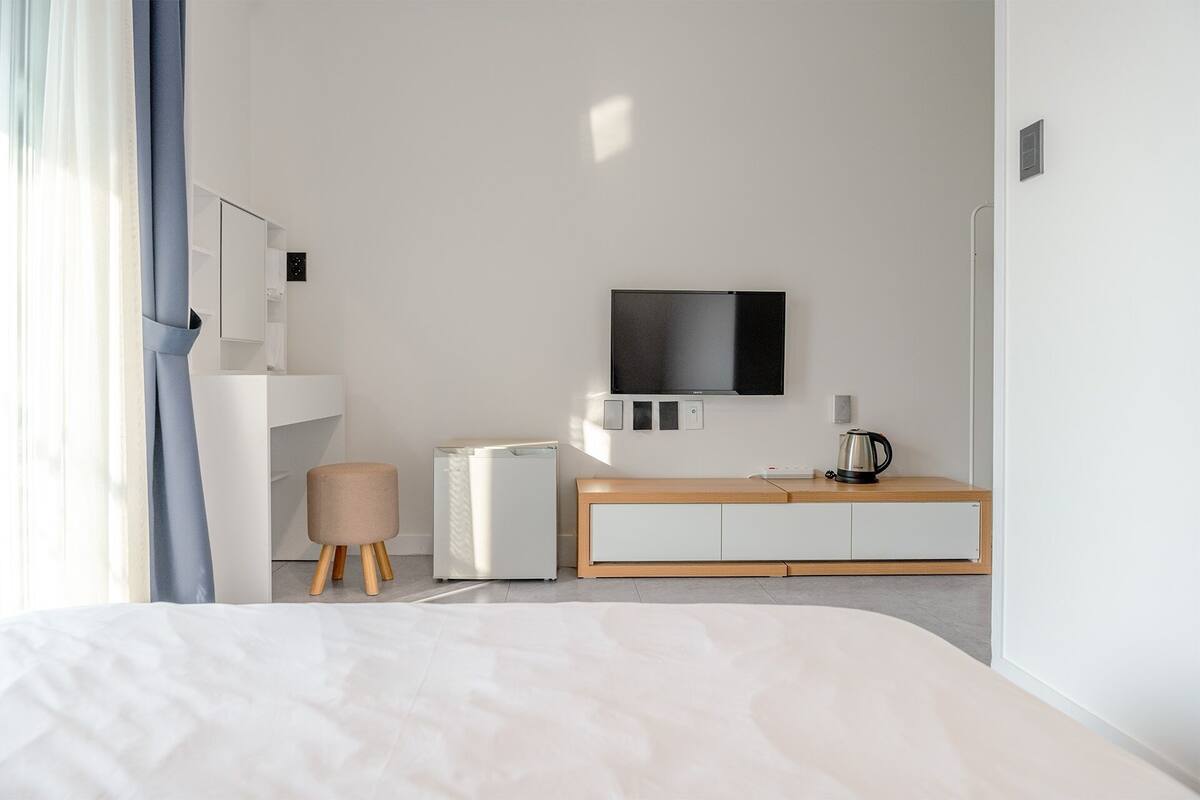 Una habitación de hotel moderna con una cama, un televisor montado en la pared, una pequeña kitchenette y una mesita de noche de madera.