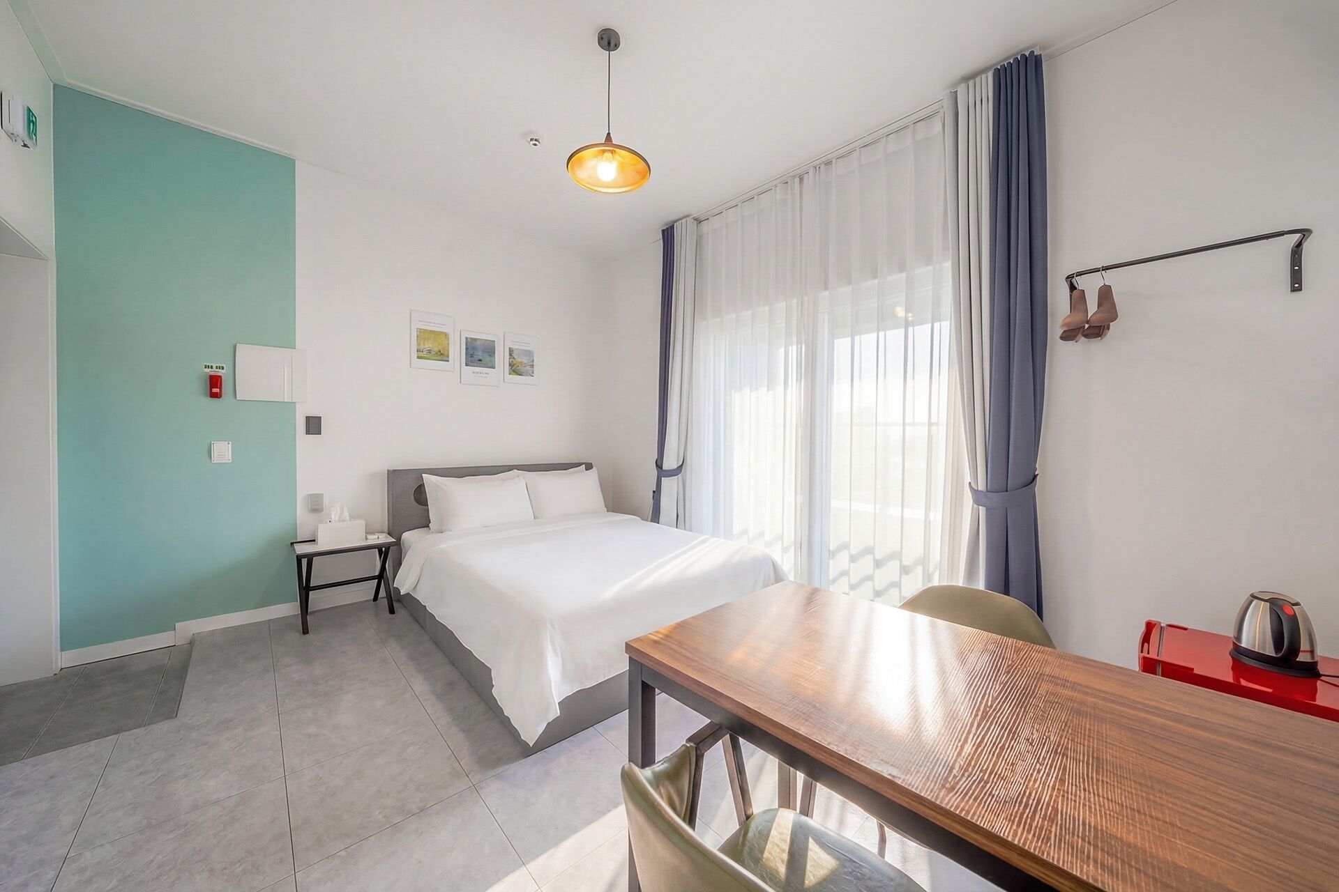 A hotel room with a bed, a table, a chair, a hanging light fixture, and a window with curtains.