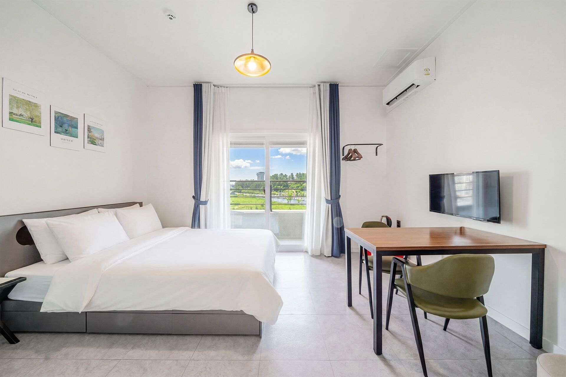 A hotel room with a bed, a desk with a chair, a television, and a view of greenery outside.