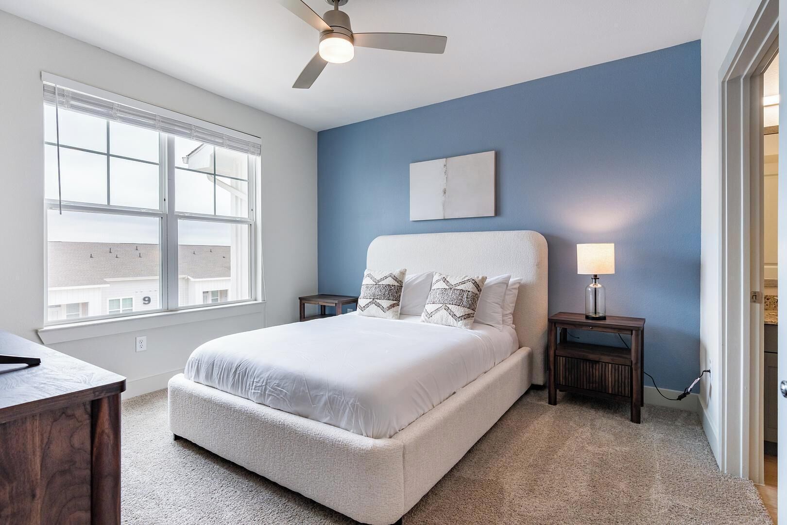 A bedroom with a bed, a ceiling fan, a nightstand with a lamp, a window, and a door.