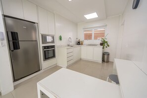Moderni keittiö, jossa on ruostumattomasta teräksestä valmistettu jääkaappi, uuni ja mikroaaltouuni, valkoiset kaapit ja ikkuna, jossa on sälekaihtimet.