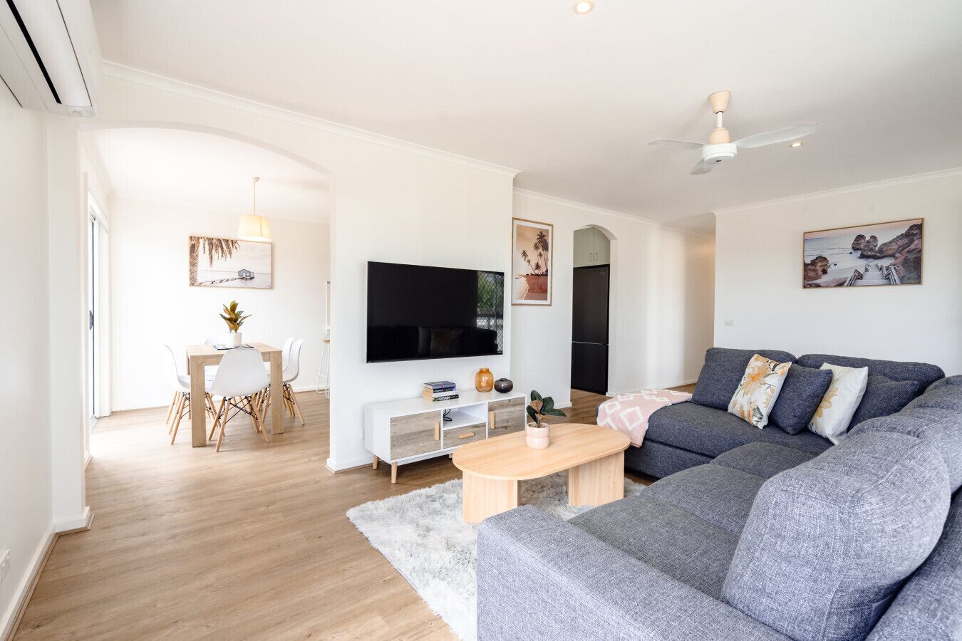 A modern living room with a grey sofa, a flat-screen TV, a dining area with a table and chairs, and a kitchen in the background.