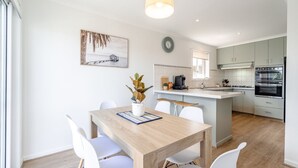 A modern kitchen with white cabinets, a central island, and a dining area with a wooden table and white chairs. - Views on the Avenue (499551) (Clifton Springs)