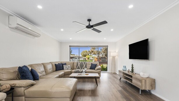 A modern living room with a sectional sofa, a coffee table, a ceiling fan, a TV mounted on the wall, and a large window with a view. - Tidal Waves on Lowe - Ocean Grove  (499548) (Ocean Grove)