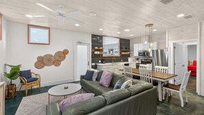 Una sala de estar moderna con un sofá verde, una mesa de comedor blanca y una zona de cocina con armarios blancos y electrodomésticos de acero inoxidable.