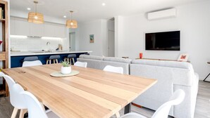 Un soggiorno moderno con tavolo da pranzo in legno, sedie bianche, divano grigio e angolo cottura con pensili bianchi e piano di lavoro blu.