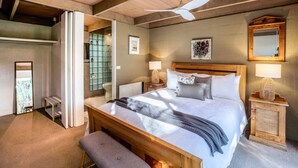 Un dormitorio con una cama de madera, un ventilador de techo, una mesita de noche con lámpara y una ventana con vistas.