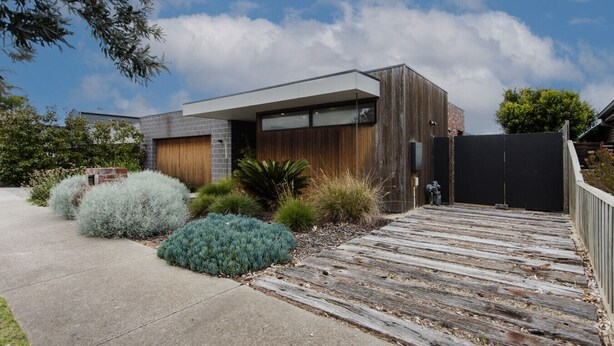 Une maison moderne dotée d’une terrasse en bois, d’un garage et d’une cour clôturée agrémentée de divers arbustes.