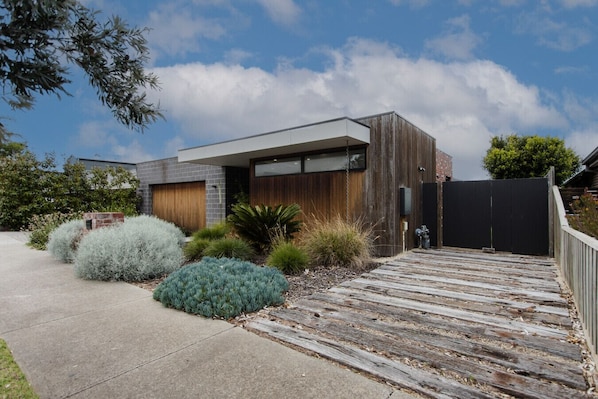 A modern house with a wooden deck, a garage, and a fenced yard with various shrubs. - Greenview Luxe - Sleeps 7 - Firepit! (499541) (Ocean Grove)