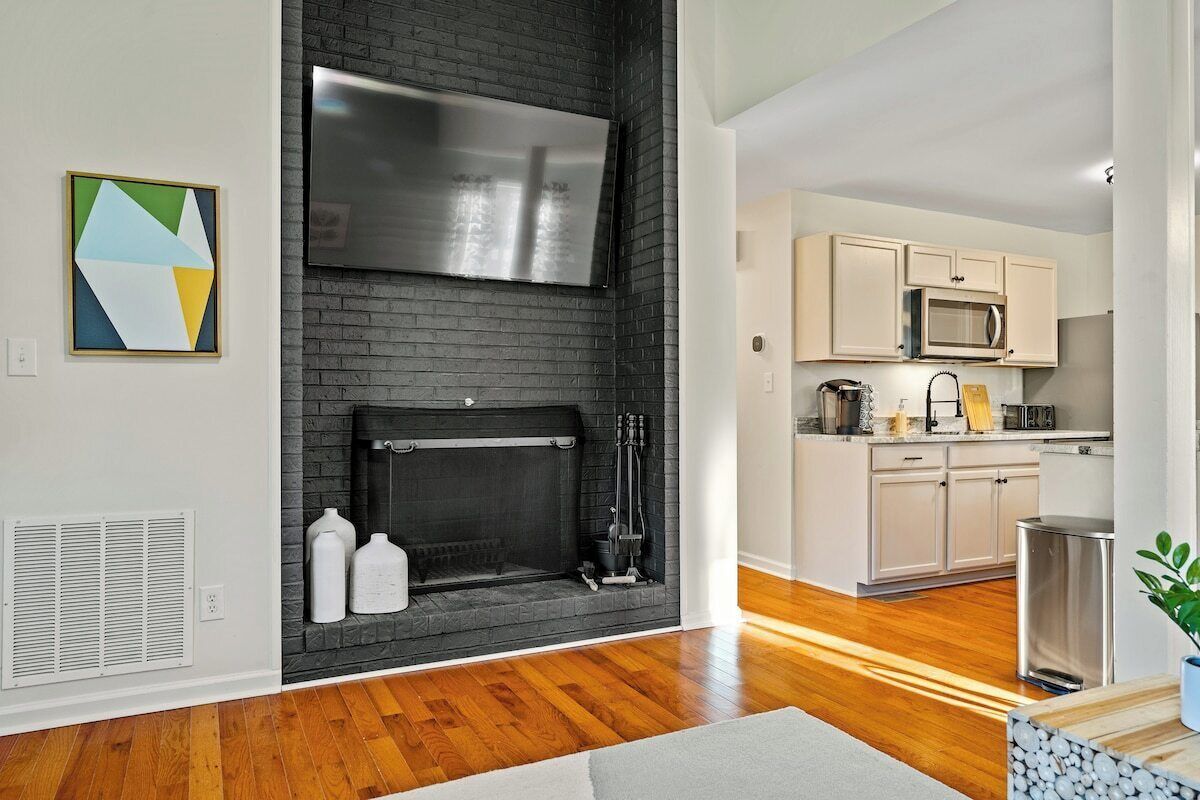A modern living room with a fireplace, a kitchen with white cabinets, and a large TV mounted above the fireplace.