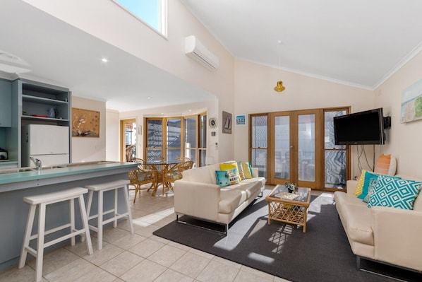 A modern living room with a sofa, coffee table, and a TV. There's a kitchen area with a fridge and a dining table.