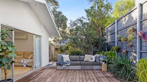 Une terrasse en bois avec un canapé et des coussins gris, entourée de plantes en pot et d’une clôture en métal.