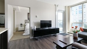 A modern living room with a flat-screen TV on a black entertainment unit, a sofa, a coffee table, and a view of the city through large windows. - Modern Spacious Waterfront Condo Downtown Calgary (Calgary)