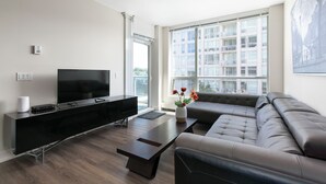 A modern living room with a flat-screen TV, a sofa, a coffee table, and a large window with a view of a building. - Modern Spacious Waterfront Condo Downtown Calgary (Calgary)