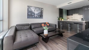 A modern living room with a sectional sofa, a coffee table with a vase of flowers, a kitchen area with dark cabinets and a bar stool, and a cityscape painting on the wall. - Modern Spacious Waterfront Condo Downtown Calgary (Calgary)
