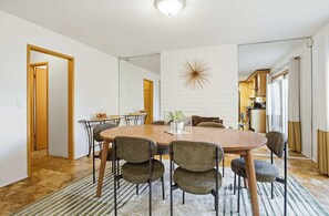 A dining area with a round wooden table, six upholstered chairs, a decorative wall piece, and a kitchen in the background. - Mid-century oasis | Private courtyard! (San Diego)