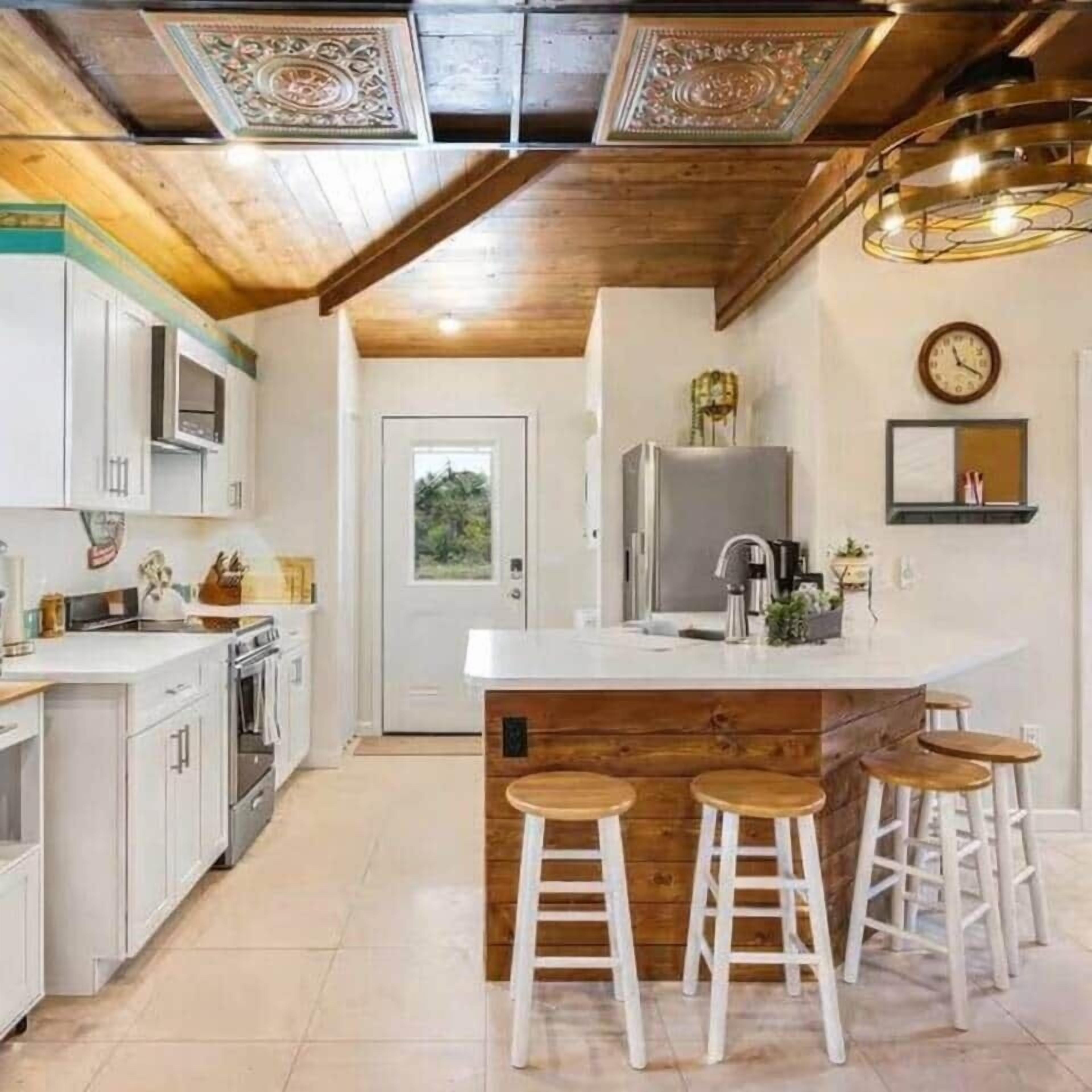 Uma cozinha com armários brancos, uma ilha de madeira com banquetas, uma geladeira e um relógio na parede.