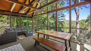 Alpendre coberto com mesa e bancos de madeira, sofá e janelas amplas que oferecem vista para árvores e colinas.