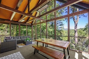 Eine überdachte Terrasse mit einem Holztisch und Bänken, einer Sitzecke und großen Fenstern mit Blick auf Bäume und Hügel.