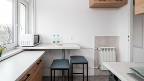 A modern kitchen with a white countertop, a microwave, two stools, and a wooden cabinet above the counter. - Limanowskiego Harmony Apartment (Łódź)