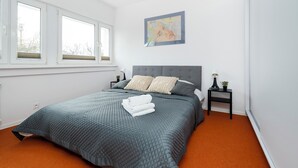 A bedroom with a bed, a window, a nightstand, a lamp, and a painting on the wall. - Limanowskiego Harmony Apartment (Łódź)