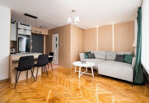 A modern living room with a sofa, a small round table, and a kitchen area with a refrigerator and cabinets. - Wola Anielewicza Deluxe Studio (Warszawa)
