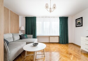 A modern living room with a sofa, a white table, a radiator, and a window with curtains. - Wola Anielewicza Deluxe Studio (Warszawa)