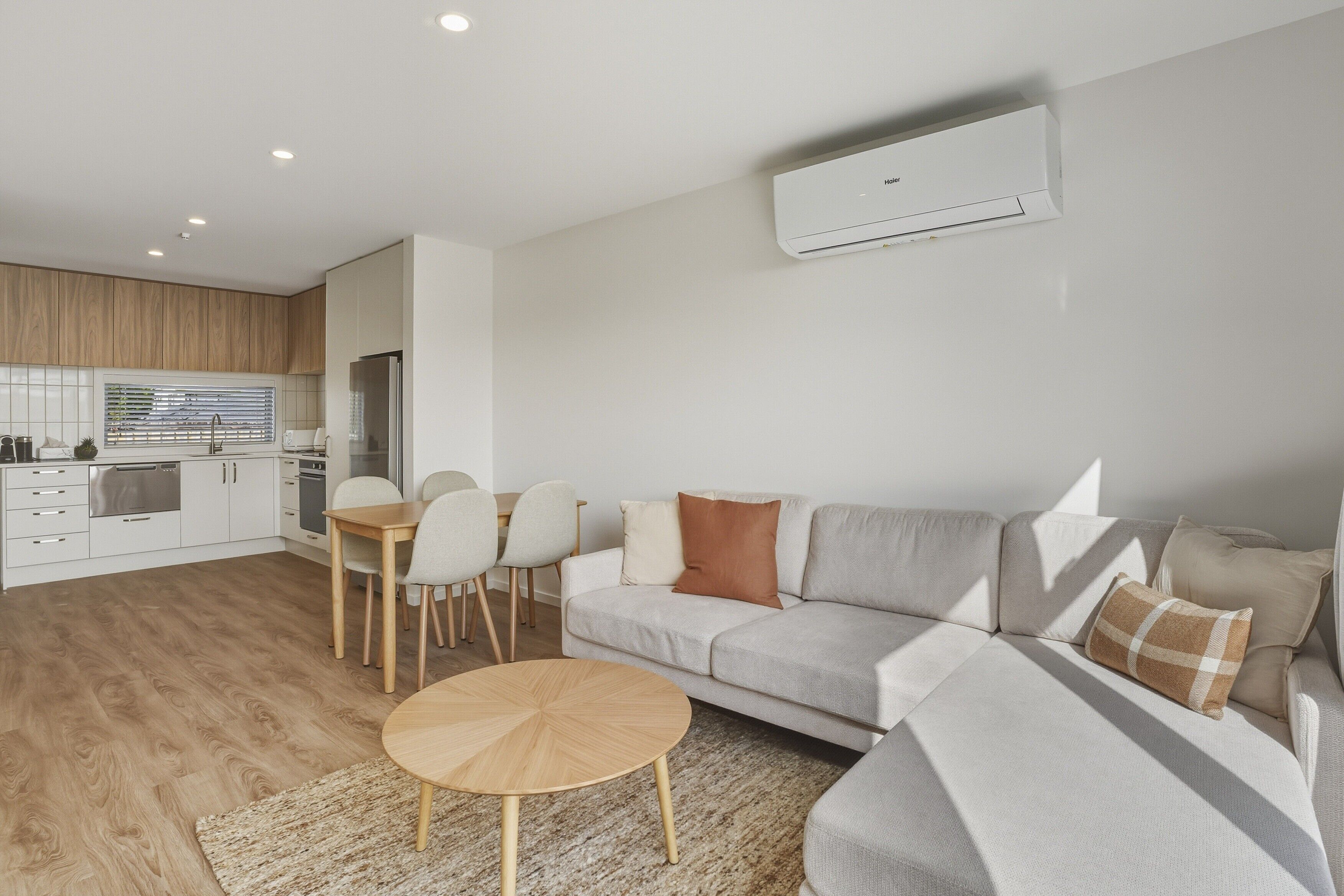 A modern living room with a sofa, a dining table, and chairs. There is a kitchen area with cabinets and a window. An air conditioning unit is mounted on the wall.