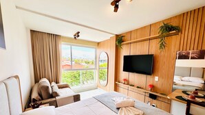 Une chambre d’hôtel avec un grand lit, un téléviseur fixé au mur, un petit bureau avec une chaise et une fenêtre donnant sur un paysage verdoyant et des bâtiments.