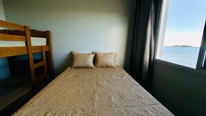 Ein Einzelbett mit gemustertem Bettbezug, zwei Kissen, einem Holz-Kopfstück und einem Fenster mit Meerblick.